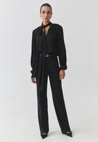 Blusa nera con collo annodato, maniche lunghe e vestibilità comoda, abbinata a pantaloni neri sartoriali e décolleté nere a punta.