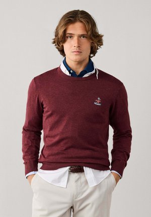 Junger Mann mit mittellangem Haar trägt einen bordeauxfarbenen Pullover über einem blauen Hemd mit Kragen und einem weißen Hemd, dazu eine helle Hose und hat die Hände in den Taschen.