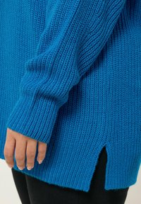 Pull en tricot bleu avec une texture côtelée, des manches longues et des fentes sur les côtés à l'ourlet, avec une coupe ample et un fil épais et doux.