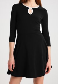 Abito nero con maniche a tre quarti, caratterizzato da un dettaglio a forma di chiave circolare sul colletto e un bordo bianco, gonna svasata.