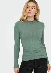 Groene ribgebreide top met lange mouwen, een ribbeltextuur en een gefronste halslijn, gecombineerd met zwarte textuurbroek.