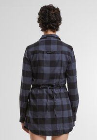 Camicia di flanella a quadri nera e blu con maniche lunghe, vita con cinturino e orlo curvo. Presenta polsini con bottoni e una spalla posteriore.