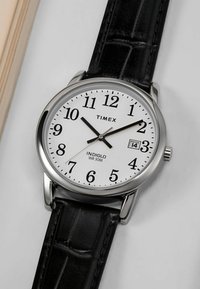 Montre Timex avec un cadran blanc, des chiffres noirs et un boîtier argenté. Dotée d'un bracelet en cuir noir avec un effet crocodile et d'un affichage de la date.