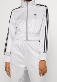 Vit zip-up träningsdräkt med svarta ränder längs ärmarna; ribbade muddar och midjeband; slätt, glansigt material; logotyp på bröstet.