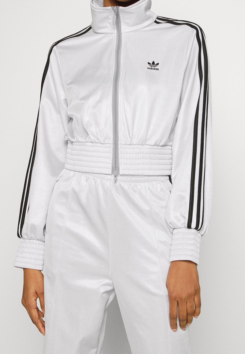 Vit zip-up träningsdräkt med svarta ränder längs ärmarna; ribbade muddar och midjeband; slätt, glansigt material; logotyp på bröstet.