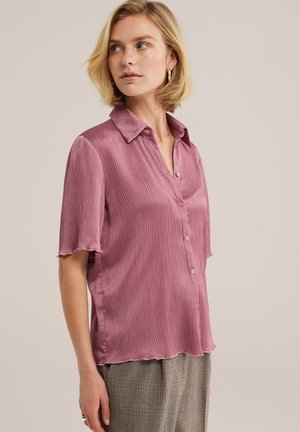 Femme portant un chemisier mauve à manches courtes boutonné avec une texture côtelée subtile et un pantalon gris, tournée vers la gauche avec des cheveux blonds courts.
