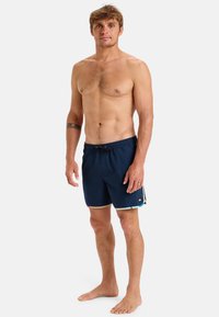 Bañador azul marino con cintura elástica, cordón y ribete en contraste. Presenta elegantes acentos laterales y un pequeño emblema de logo.