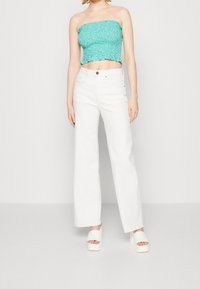 Tyrkysový květinový top bez ramínek s volánkovými okraji, doplněný širokými bílými džíny. Modelka nosí bílé platformové sandály, čímž předvádí uvolněný vzhled.