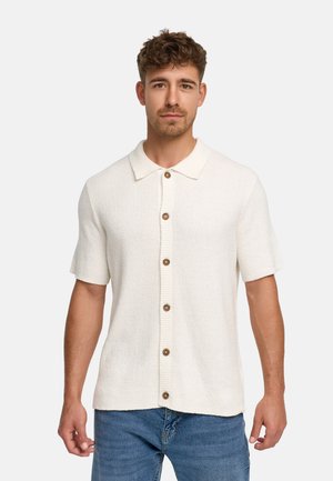Junger Mann mit kurzen lockigen Haaren, der ein cremefarbenes kurzärmliges Strickhemd mit Knöpfen und blaue Jeans trägt und vor einem schlichten Hintergrund steht.