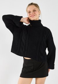 Pull en tricot noir avec un col montant, présentant des motifs texturés et un design cropped. Associé à une jupe noire.