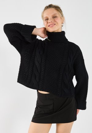 Black knitted sweater with a high collar, featuring textured patterns and a cropped design. Paired with a black skirt.
