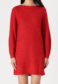 Una persona che indossa un vestito lungo a maniche, lavorato a maglia, di colore rosso si trova davanti a uno sfondo bianco uniforme, mostrando la texture del tessuto.