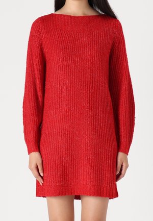 Une personne portant une robe rouge en tricot à manches longues se tient devant un fond blanc uni, mettant en valeur la texture du tissu.