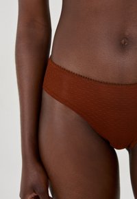 Brauner Bikini mit Punkten, weicher Textur und gewelltem Rand. Die Seitenansicht konzentriert sich auf den Taillenbund und den glatten Stoff auf der Haut.