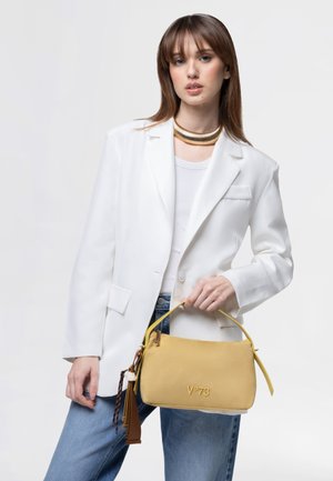 Giovane donna con blazer bianco e jeans blu tiene una borsa gialla V°73 con dettagli a frange su uno sfondo semplice.