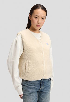 Donna che indossa un gilet in pile color crema sopra una camicia bianca a maniche lunghe e jeans blu, in piedi davanti a uno sfondo grigio uniforme.