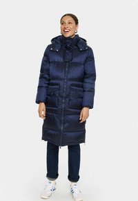 Tumši zilo pūkaini mēteli ar kapuci, ar rāvējslēdzēju, priekšējām kabatām un quilted dizainu. Saskaņots ar tumšajām džinsiem un baltajām kedām.