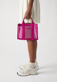 Transparent plastväska med livfulla rosa detaljer, stadiga handtag och ett texturerat mönster på botten, visad bredvid en person i sneakers.