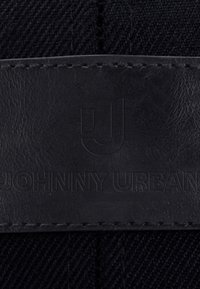 Zwart leren patch genaaid op donkere stof met geperst tekst "JOHNNY URBAN" en een gestileerd "JU" logo boven de tekst.