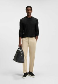 Svart texturerad tröja, beige dressbyxor, svarta sneakers. Håller en svart läderväska med rundade kanter. Enkel, modern design.