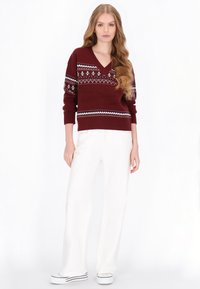 Bordowy sweter z dzianiny z dekoltem w kształcie litery V i geometrycznymi wzorami w bieli i niebieskim. W połączeniu z białymi spodniami o szerokich nogawkach i casualowymi sneakersami.