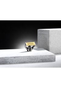 Sähköinen partakone kromisella pohjalla ja kullanvärisellä kalvopäällä, asetettuna harmaalle betonipinnalle tumman taustan edessä.