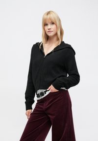 Hanorac negru scurt, cu fermoar în față, cu manșete și tiv din material striat. Purtați peste un top alb cu volane și asortat cu pantaloni burgundy din catifea cu dungi.