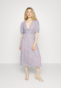 Vestido com estampado floral em rosa e azul pastéis, com decote em V profundo, mangas bufantes e comprimento midi fluído. Usado com sapatos de salto bege com tiras.