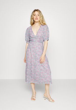 Vestido com estampado floral em rosa e azul pastéis, com decote em V profundo, mangas bufantes e comprimento midi fluído. Usado com sapatos de salto bege com tiras.