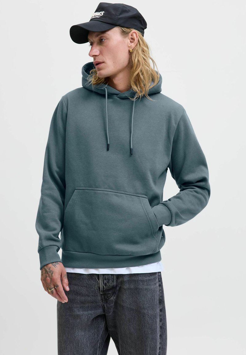 Türaquahi-Hoodie mit Kordelzügen, Fronttasche und gerippten Bündchen. Trägt dazu graue Jeans und eine schwarze Kapuze mit einem Logo auf der Vorderseite.