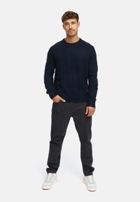 Marineblauw wollen mengsel crew-neck trui met geribbelde manchetten en onderrand, gecombineerd met zwarte denim jeans en witte sneakers, staand tegen een effen achtergrond.