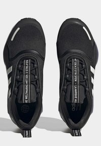 Ett par svarta träningsskor med vita ränder, slip-on stil, elastiska snörningar och märkta dragflikar med flerspråkig text på tungan.