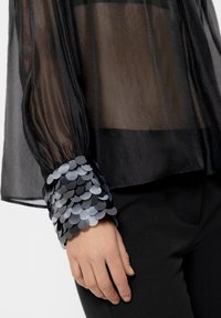 Schwarze, durchsichtige Bluse mit puffärmel. Die Manschetten sind mit überlappenden grauen und silbernen, runden Pailletten verziert. Kombiniert mit schwarzen Hosen.