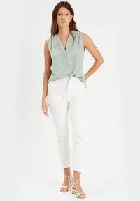 Chemise sans manches vert clair avec un col en V et des sequins éparpillés, associée à un jean blanc ajusté et des sandales à talons blocs dorés.