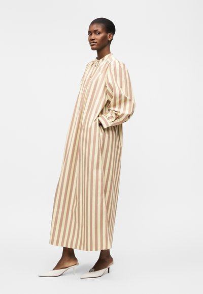 Γυναίκα όρθια που φοράει μακρύ μπεζ και ανοιχτό καφέ φόρεμα με κάθετες ρίγες και μακριά μανίκια, καθώς και λευκές ψηλοτάκουνες mule, με τα χέρια στις τσέπες.