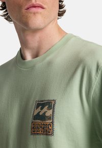 Ανοιχτό πράσινο βαμβακερό μπλουζάκι με στρογγυλή λαιμόκοψη, με γραφικό σχέδιο κυμάτων και το κείμενο "BILLABONG" σε σκούρο μπλε και πορτοκαλί.