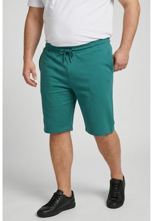 Uomo che indossa pantaloncini turchese con coulisse, t-shirt bianca e scarpe da ginnastica nere, in piedi davanti a uno sfondo semplice.