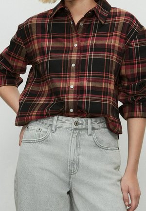 Personne portant une chemise à carreaux aux manches retroussées dans des tons rouges, noirs et marron, associée à un jean taille haute gris clair.
