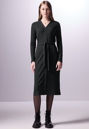 Zwarte geribbelde jurk met een V-hals, lange mouwen en een gestrikt taille. Heeft knopen aan de voorkant en een middenlange snit, in combinatie met zwarte veterschoenen.
