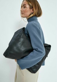 Divas melnas ādas somas, viena ar tekstūru un liela, turētas zem rokas. Cilvēks ir ģērbies gaiši zilā džemperī un krēmkrāsas biksēs.