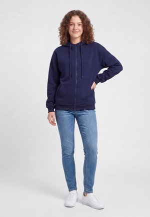 Tânără cu păr creț, purtând un hanorac bleumarin cu fermoar, blugi bleu și pantofi sport albi, stând cu o mână pe șold.