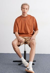 Rustovo sfarbené oversized tričko, béžové kraťasy, sediace na stoličke. Biele tenisky s svetlohnedými akcentmi. Krátke, kučeravé vlasy.