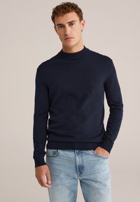 Man med lockigt hår som bär en marinblå långärmad tröja och ljusblå jeans, står med ena handen i fickan.