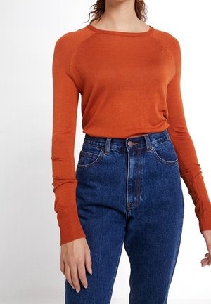 Femme portant un pull ajusté à manches longues orange brûlé rentré dans un jean taille haute bleu foncé, sur un fond blanc.