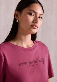 Mladá žena s dlouhými tmavými vlasy, která má na sobě naušnice a růžové tričko s nápisem "new york city", na neutrálním pozadí.