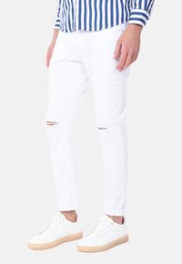 Jeans in denim bianco con vestibilità slim, caratterizzati da tagli sfrangiati sulle ginocchia. Abbinati a sneakers bianche con suola beige chiaro. Camicia a righe blu e bianche visibile.