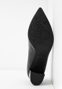 Chaussure à talon noir avec un bout pointu, tige en cuir lisse et semelle en caoutchouc texturée avec des détails embossés et du branding.