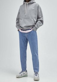 Felpa grigia con tasca frontale, cordini e logo. Sovrapposta a una maglietta a righe rosa e blu navy. Jeans azzurri e sneakers grigie con dettagli.