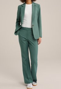 Grön skräddarsydd blazer och matchande byxor, tillverkade av mjukt tyg. Bärs över en vit skjorta, kompletterad med vita sneakers.