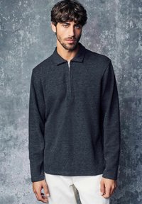 Tamno sivi polo s dugim rukavima i polu-zipper izrezom. Tekstura tkanine pokazuje suptilan uzorak. Model nosi bijele hlače.